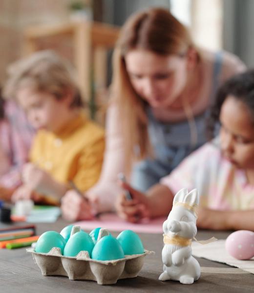 Grey and white toy rabbits and group of painted Easter eggs in egg-box standing on wooden table with kids drawing pictures on background